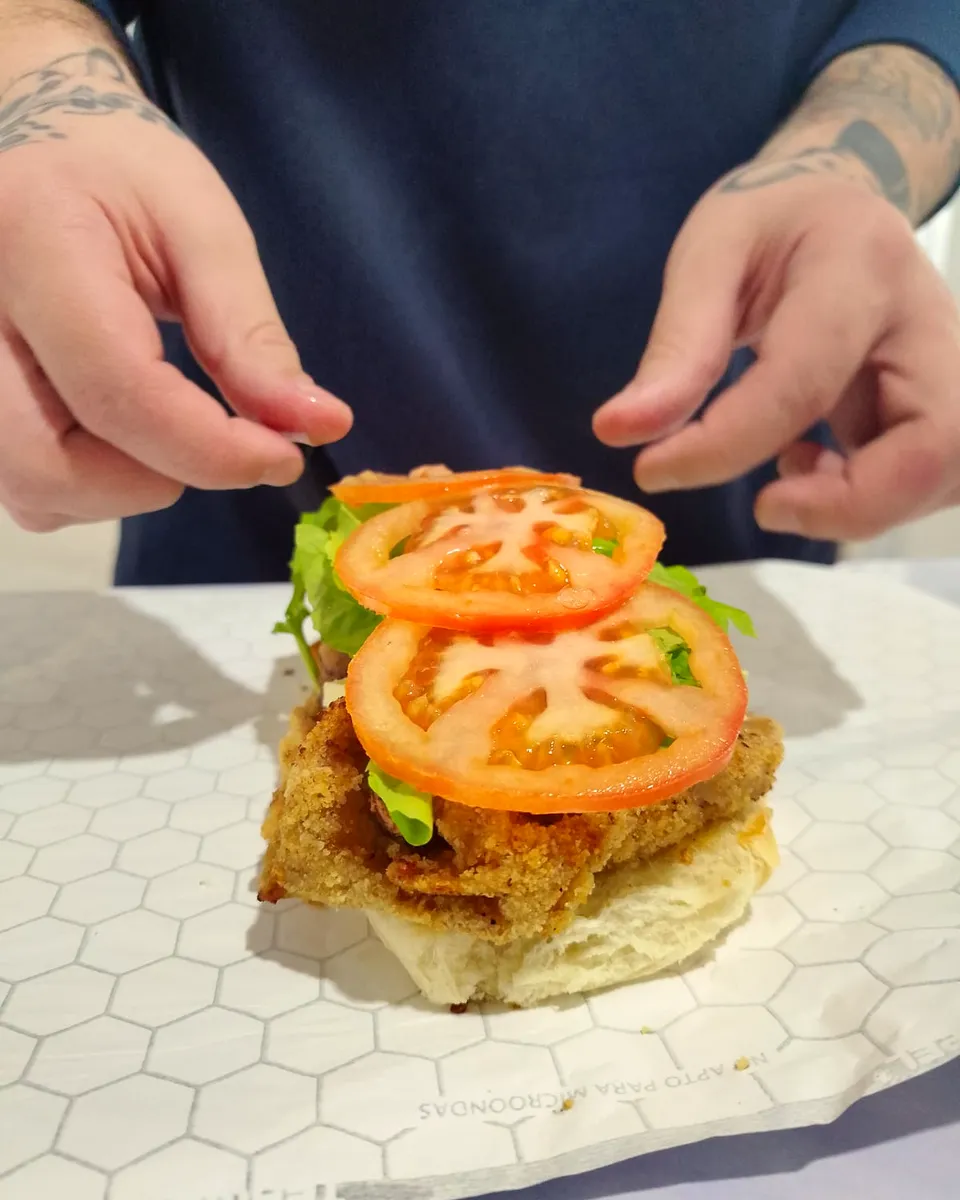 Manos armando un sanguche: milanesa, lechuga y rodajas de tomate sobre el pan